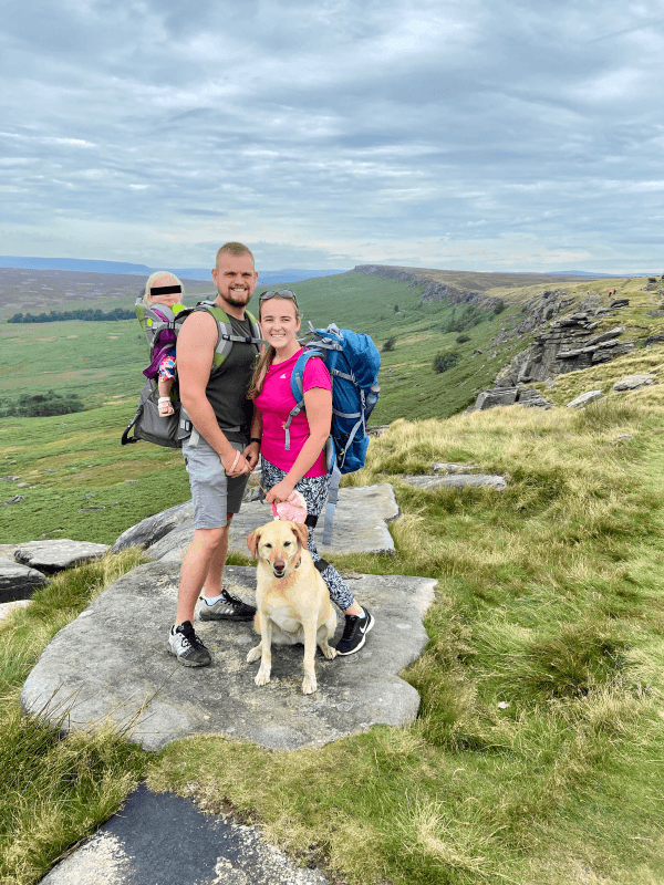 Rhiannon, Luke, Mollie & Daisy - @thehappycamper2020 10/2020 A smiling family of three with a baby in a carrier and a golden retriever dog, standing on a rocky outcrop during a hike in the UK countryside, with rolling hills in the background.