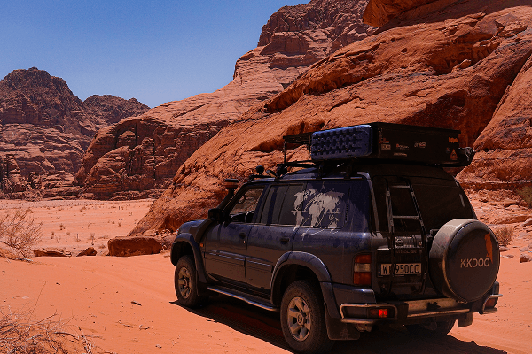 Long days of discovering canyons, rock formations, and mountains - @kkdoo_pl 03/2023 The blue Nissan Patrol Y61 overland 4x4 camper of kkdoo_pl is parked on the red sands of Wadi Rum, Jordan, surrounded by towering sandstone cliffs and desert canyons. The vehicle is equipped with a rooftop tent and travel gear, symbolizing Kasia and Constantin’s vanlife adventure and exploration through the Middle East.