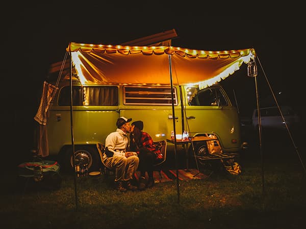 Anna & Christoph in love with Heinrich - @meetheinrich 06/2022 A couple, Anna and Christoph from @meetheinrich, enjoying a cozy evening outside their vintage VW T2 bus, Heinrich, under a lit awning during a night camping adventure. The warm glow of string lights adds to the relaxing atmosphere of their vanlife experience.