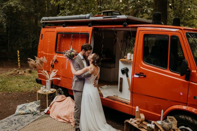 Our van wedding in the woods - @the.redcamper 10/2020 A newlywed couple sharing a tender moment beside a vibrant orange vintage van. The bride, in a flowing white gown, and the groom, in a smart grey suit, are embracing and gazing into each other’s eyes. The van’s door is open, revealing a cozy interior, and the scene is adorned with rustic decorations including a string of lights, blankets, pillows, and a makeshift wooden shelf with various personal items, set in a tranquil outdoor setting with trees in the background. @the.redcamper 10/2020 - Vanlifezone (Issue 2)