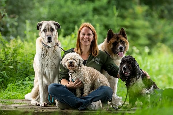 Dogs and humans are more alike than a lot of people know. - @lizwolting 12/2024 Liz Wolting (@lizwolting), an animal behaviorist and advocate for dogs, sits outdoors on a wooden platform surrounded by lush greenery, accompanied by four diverse dogs, including a large Anatolian Shepherd, a fluffy curly-haired dog, an Akita, and a Spaniel. Smiling warmly, Liz showcases her bond with the dogs, reflecting her mission to help canines with behavioral challenges while embracing her outdoor lifestyle.