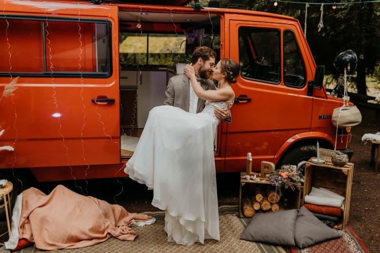 Van-wedding fairy tale - @the.redcamper 10/2020 A newlywed couple sharing a tender moment beside a vibrant orange vintage van. The bride, in a flowing white gown, and the groom, in a smart grey suit, are embracing and gazing into each other’s eyes. The van’s door is open, revealing a cozy interior, and the scene is adorned with rustic decorations including a string of lights, blankets, pillows, and a makeshift wooden shelf with various personal items, set in a tranquil outdoor setting with trees in the background. @the.redcamper 10/2020 - Vanlifezone (Issue 2)