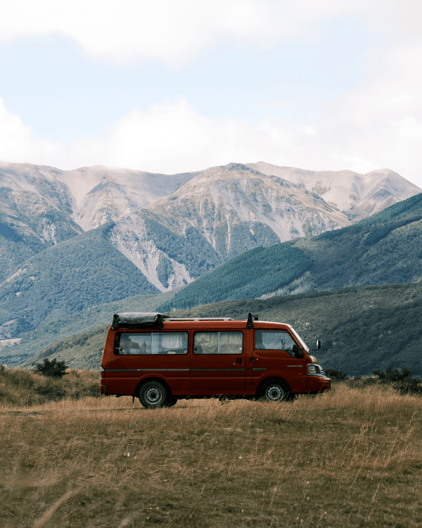 Four wheels and two cameras - @carnet_declaireurs 06/2024 Red camper van parked in a scenic mountainous area of New Zealand, showcasing the vanlife experience. The vehicle is set against a backdrop of lush green hills and rugged mountains under a partly cloudy sky, emphasizing the adventurous spirit of travel photographers exploring the natural beauty of New Zealand.