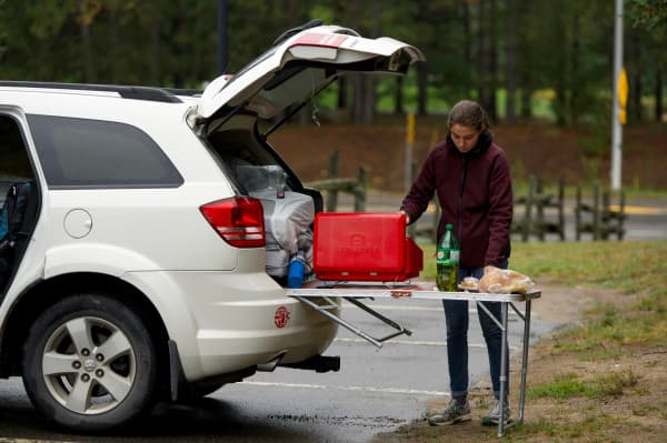 Claire preparing a meal at the back of their car in Canada - @carnet_declaireurs 06/2024 Claire from @carnet_declaireurs setting up a camping stove and preparing a meal at the back of a white SUV during a camping adventure in Canada. The vehicle's trunk is open, revealing camping gear and supplies, capturing the essence of mobile travel and outdoor cooking in a parking lot in Canada.