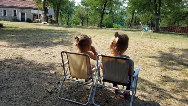 Sister time during van life - @feel_free_van 02/2022 Two young girls, Stella and Zara, sit side by side in small camping chairs in a sunny, rural yard in Bulgaria. They are enjoying a quiet moment together, surrounded by trees and a rustic stone house in the background. The scene captures a peaceful and simple moment of childhood during their family's van life adventures.