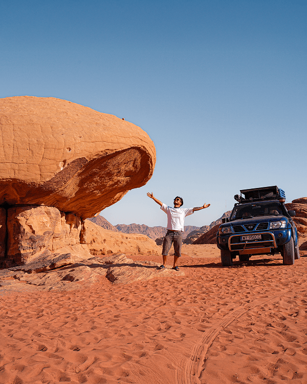 Exploring the red desert of Wadi Rum - @kkdoo_pl 03/2023 Constantin from kkdoo_pl stands with open arms beside their blue Nissan Patrol Y61 overland 4x4 camper in the red desert of Wadi Rum, Jordan. Towering sandstone rock formations and clear blue sky frame the scene, capturing the spirit of adventure, vanlife, and overlanding through the Middle East.