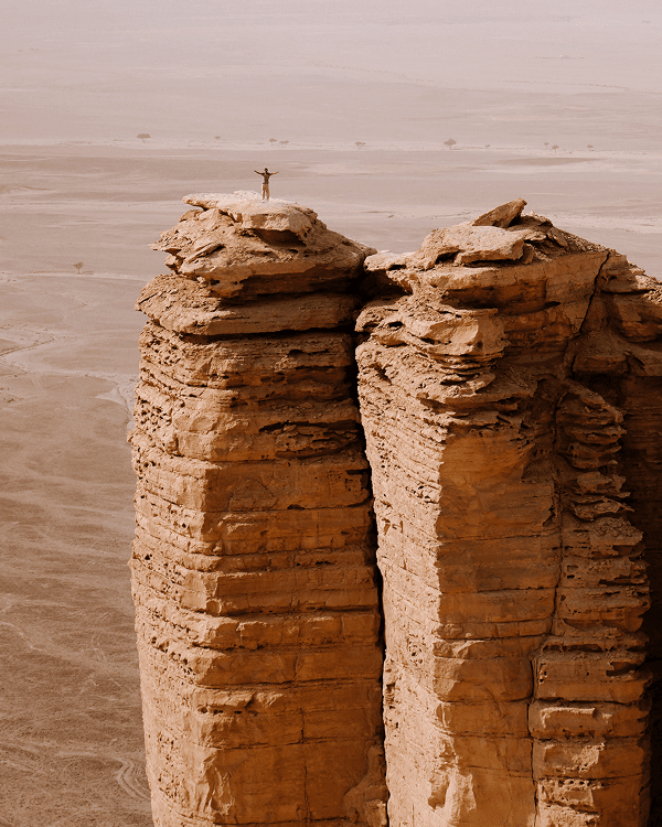 The desert was alive and varied - @countless_roads 03/2023 One of the travelers from @countless_roads stands triumphantly on top of a towering sandstone pillar overlooking the vast desert plains of Saudi Arabia. The scene captures the raw scale and beauty of the Arabian landscape, symbolizing freedom, adventure, and the spirit of overlanding experienced by Amy and Jeroen during their vanlife journey across the Arabian Peninsula.