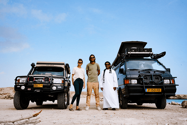 An Omani overlander with the dream of driving to Africa - @countless_roads 03/2023 Amy and Jeroen from @countless_roads stand beside their Mitsubishi L300 4x4 with an Omani overlander and his Land Cruiser, both vehicles parked on a rocky desert plain under a bright blue sky. The image captures the spirit of cross-cultural connection, overlanding friendship, and vanlife exploration across the Arabian Peninsula.