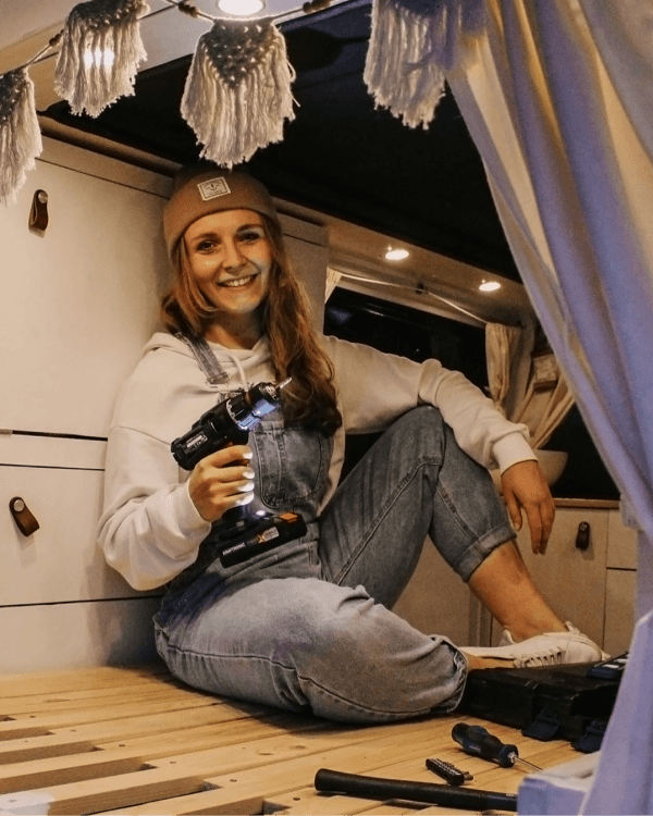 Jenni in her self-built camper van - @itsme.jenni 02/2022 A woman (@itsme.jenni) sitting inside her self-built camper van, holding a power drill. She is smiling and appears proud of her work. The van interior includes wooden furniture and cozy decor, reflecting her journey of transforming a VW T5 into a mobile home.