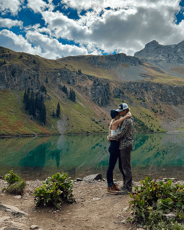 Going on tough hikes in the national parks - @wanderingnwonder 06/2025 Peter and Tara of @wanderingnwonder embrace beside a turquoise alpine lake surrounded by mountains and evergreen trees under a partly cloudy sky. Captured during their vanlife journey through the USA, this moment reflects their deep connection, love for hiking, and appreciation for wild and remote landscapes.