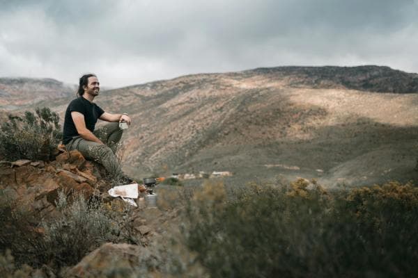 Stay hydrated on your adventures - Pexels/Taryn Elliott A man sits on a rocky outcrop in the mountains, enjoying a break during his outdoor adventure. He is dressed in casual hiking attire with a black t-shirt and khaki pants, holding a cup of what appears to be a hot beverage. The landscape behind him features rolling hills and a cloudy sky, creating a serene and picturesque scene. Camping equipment, including a portable stove, can be seen beside him, indicating a well-prepared hiking or camping trip. This image captures the essence of outdoor exploration and the tranquility of nature.