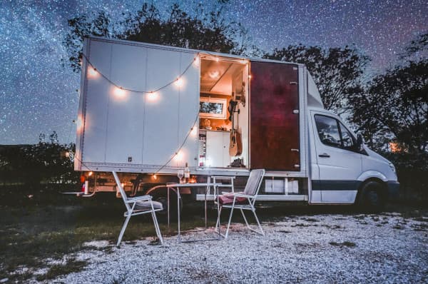 Parked up under the stars - @offthemainroad_ 08/2021 Cozy mobile tiny home converted from a white cargo van parked under a starry night sky, featuring an open back with string lights, a comfortable interior setup visible through the back doors, and outdoor seating area with two white chairs and a small table on a gravel surface.