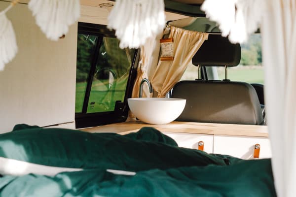 Even though mistakes happened, the interior looks fabulous - @itsme.jenni 02/2022 The interior of a self-built VW T5 camper van, featuring a neatly arranged sink, wooden countertop, and cozy green bedding. The van, transformed by Jenni (@itsme.jenni), showcases her DIY skills and attention to detail.