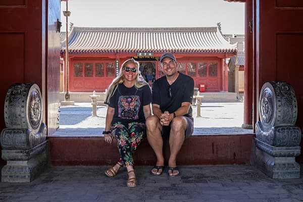 Charelle & Marcel in China - @char.is.far 01/2025 Charelle and Marcel from @char.is.far sitting together at a historic section of the Great Wall of China. The image features traditional Chinese architecture in the background, showcasing red walls and tiled roofs. This photo highlights a key moment in their overlanding adventure from Europe to Asia, emphasizing their journey through iconic landmarks in China.