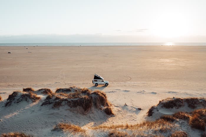 Rømø beach, Denmark - @hypespeed 09/2022 @hypespeed's white Land Rover Defender with a raised pop-up roof tent is parked on an expansive sandy beach, surrounded by a vast, open landscape. The vehicle sits near a small dune formation with patches of dry grass, contrasting against the smooth, endless sand. The golden light of the setting sun casts long shadows, highlighting the serene and remote atmosphere. The horizon stretches out with the ocean faintly visible in the background, creating a perfect scene of solitude and adventure. This image encapsulates the essence of overlanding, van life, and the freedom of remote work in nature.