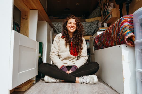 Georgia in her van - @georgiabroderick 08/2021 Georgia Broderick sits cross-legged and smiling inside her van, showcasing the efficient use of space with visible storage solutions. The van's cozy interior includes a colorful knitted blanket and an assortment of personal items, capturing a moment of joy in the compact living area of her home on wheels, ideal for those interested in van life and travel.
