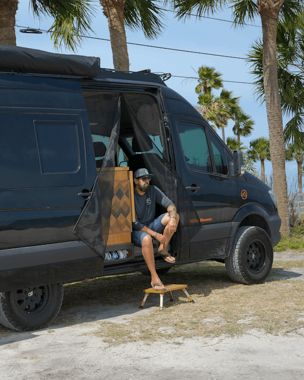 Gregg Pappas in the sliding door of his Mercedes Sprinter - @deafroamer 06/2025 Gregg Pappas, a deaf vanlifer and founder of @deafroamer, sits in the sliding door of his black Mercedes Sprinter van, enjoying a quiet moment under the palm trees near a coastal campsite in Florida. Wearing flip-flops, a cap, and sunglasses, Gregg rests on a small yellow step stool as the van’s custom woodwork and minimalist off-grid setup are partially visible behind a mesh screen. The scene captures his commitment to accessible travel and inclusive vanlife, reflecting a lifestyle built on connection, self-reliance, and community visibility for the deaf and hard of hearing.