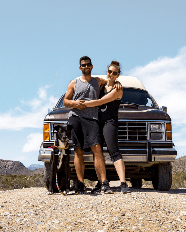 Hayley, Jake & Finnley in Texas - @hayleyandjake 10/2020 A happy couple with their dog standing in front of a classic 1985 Dodge B250 van on a dirt road in Texas, with clear blue skies and desert landscape in the background.