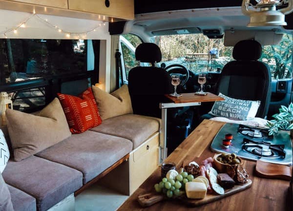 A cozy interior - @emsvanlife 08/2021 Cozy interior of Emily Jay's RAM Promaster, featuring a comfortable seating area with cushions, string lights, and a small dining setup. The wooden countertop has a charcuterie board with various snacks, and a portable stove. The inviting space showcases the practical and charming aspects of vanlife, set against a backdrop of the outdoors in Canada.
