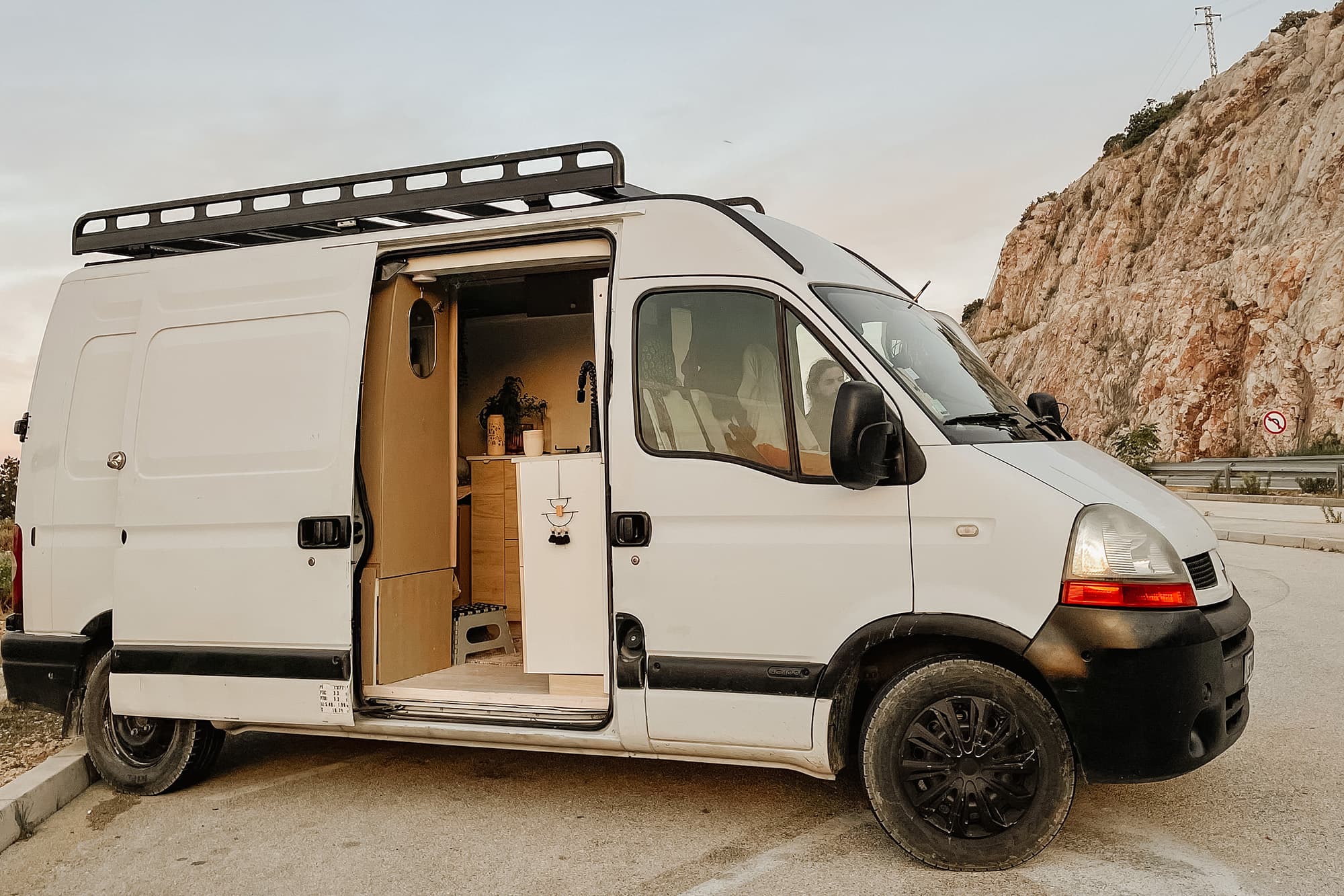 Hi, I'm Maki! - @irisandmax 06/2022 The converted Renault Master by @irisandmax van parked with its side door open, showcasing a cozy and functional interior with a kitchen area. The van is set against a backdrop of rocky cliffs and an open sky, perfect for vanlife and off-grid adventures.