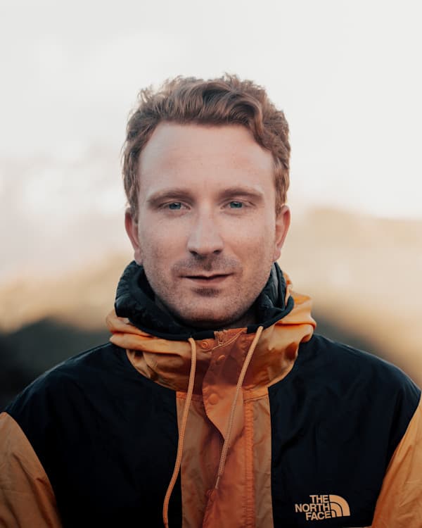 Hi, I’m Lucien - @hypespeed 09/2022 A close-up portrait of Lucien Marti (@hypespeed), an outdoor enthusiast, captured in soft natural lighting. He is wearing a two-toned black and orange The North Face windbreaker with the hood down, revealing short, slightly tousled, reddish-blond hair. His expression is calm and confident, with blue eyes gazing directly at the camera. The blurred background suggests an outdoor setting, possibly in a mountainous or rugged environment, reinforcing an adventurous and nomadic lifestyle. The warm color tones and soft focus create a natural and serene atmosphere, aligning with the theme of van life and remote work.