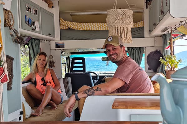 Renée & Joris inside their camper "Otto" - @travelcrazy.nl 09/2022 Renée and Joris from @travelcrazy.nl inside their camper van Otto, with a macrame decoration hanging from the ceiling, customized storage cabinets, a cozy bed area, and a view of the ocean through the front windshield, highlighting their unique and personal van interior.