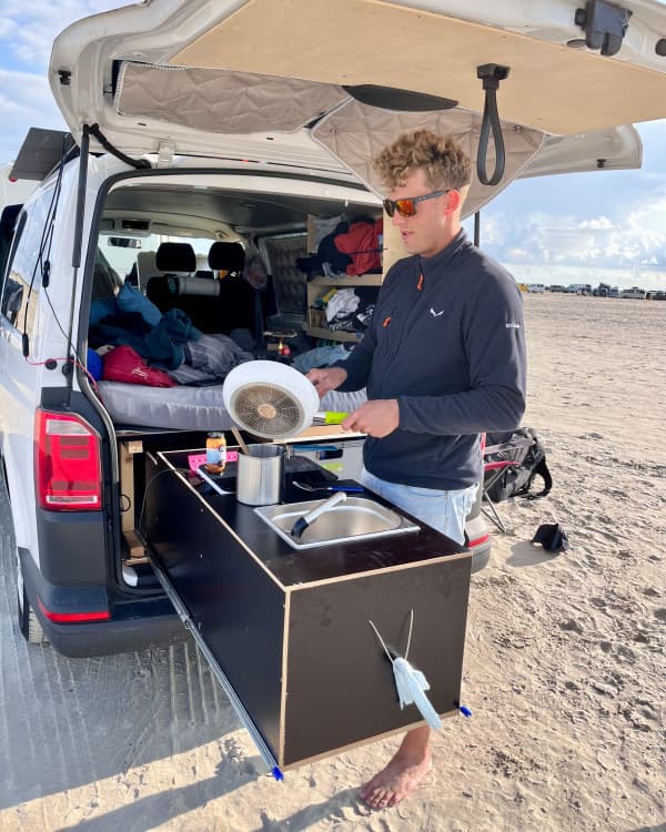 Hey everyone, I'm Chris - @van4life__ 10/2024 Chris, @van4life__ cooking outdoors at Romo Beach using the custom kitchen setup in his DIY-converted VW T6 camper. The camper's rear hatch is open, showcasing a compact and functional interior with a bed and storage. The sandy beach setting emphasizes the freedom and flexibility of vanlife.