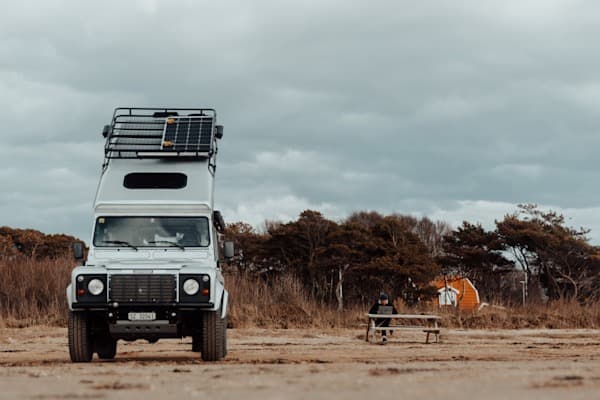 Every office on the road is different - @hypespeed 09/2022 @hypespeed's white Land Rover Defender with an extended pop-up roof tent and mounted solar panels is parked on a sandy surface under a cloudy sky. The rugged overlanding vehicle, equipped for off-grid living, stands in the foreground, while a person dressed in dark clothing sits at a wooden picnic table in the distance, possibly working or enjoying a break. Behind them, a row of wind-swept trees and a small orange tent add depth to the remote outdoor setting. The scene captures the essence of van life, self-sufficiency, and digital nomadism in a wild, natural environment.