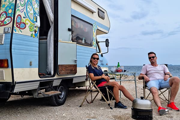 Relaxing by the Aegean Sea - @travelcrazy.nl 09/2022 Renée and Joris relaxing outside their colorful camper van Otto near Kavala, Greece, seated by a table with a scenic coastal backdrop and the Aegean Sea in view.