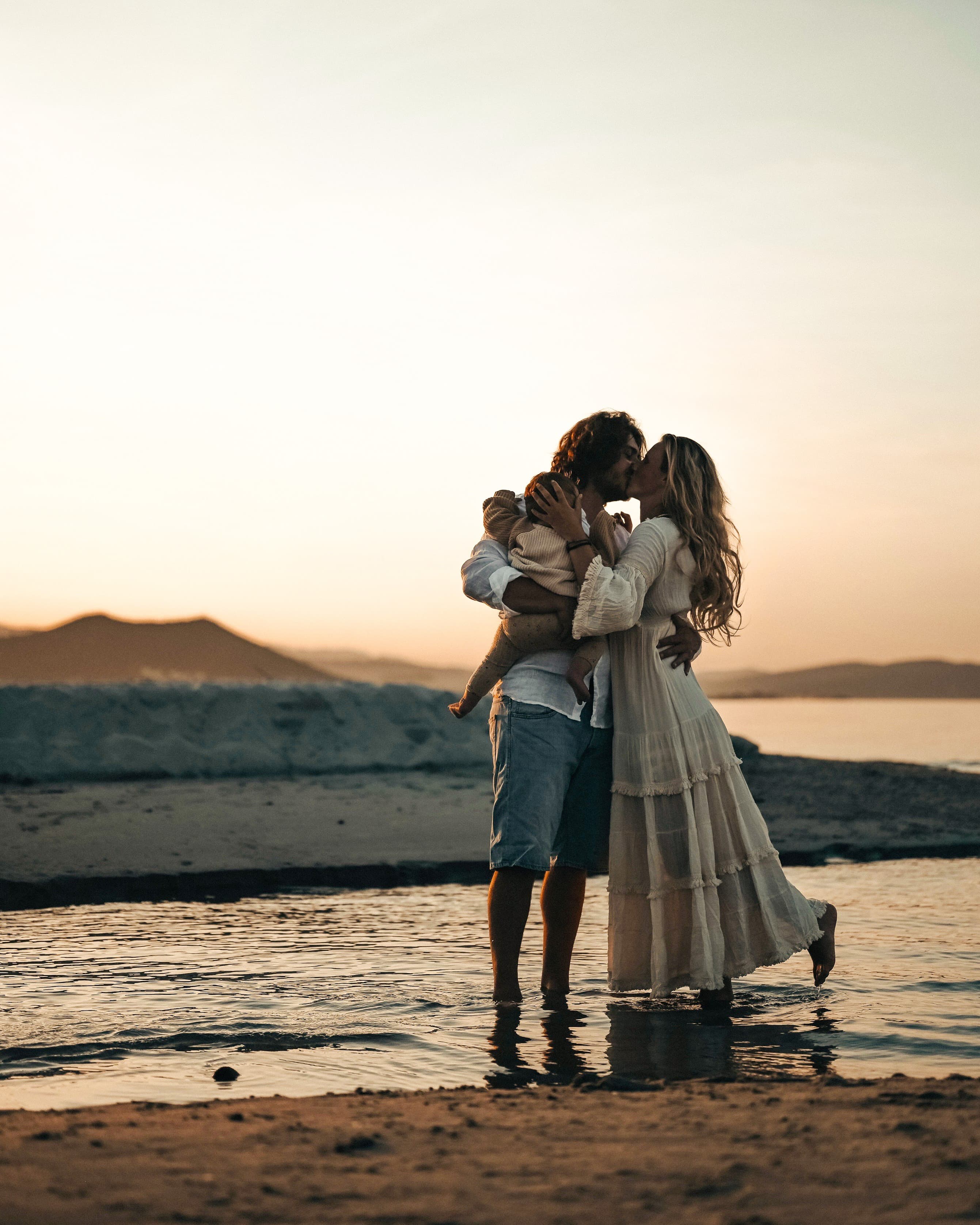 Steffi & Pascal with their baby - @h.i.p.p.i.e_l.i.f.e 09/2022 Steffi and Pascal (@h.i.p.p.i.e_l.i.f.e) stand barefoot in shallow water at sunset, holding their baby between them as they share a kiss. Dressed in soft, natural tones, they embody the freedom of vanlife and family travel. A calm sea, distant mountains, and golden light create a warm, peaceful atmosphere.