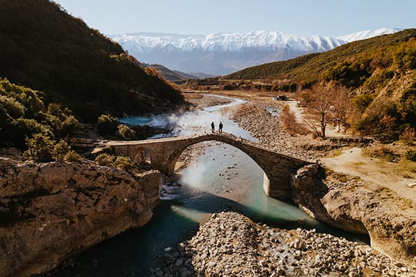 Beautiful Permet, Albania - @edelweiss.on.the.road 06/2022 Thomas & Regina, from @edelweiss.on.the.road, are seen crossing a historic stone bridge in Permet, Albania, with snow-capped mountains in the background. This scenic moment highlights their vanlife journey as they explore Albania’s natural beauty and remote landscapes.