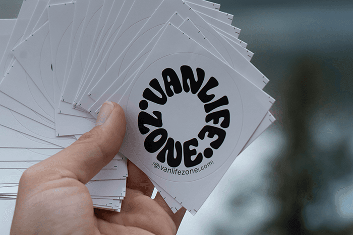 Vanlifezone stickers A hand holding a fan of circular Vanlifezone logo stickers with bold black lettering on white background, designed for travel gear and vanlife accessories.